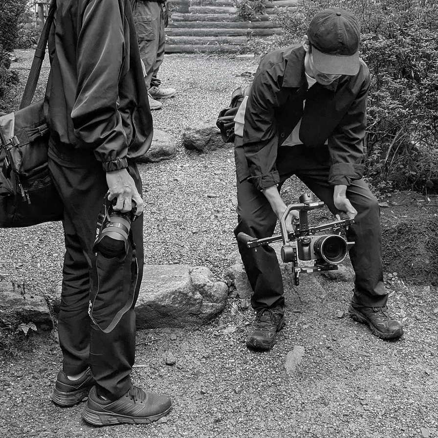 屋外でジンバルカメラを構え低アングルで撮影するスタッフ(モノクロ写真)